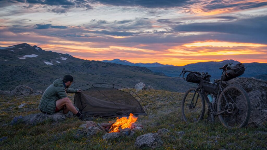 Bikepacker setting up a lightweight bivy tent beside their fully loaded gravel bike at a wild campsite