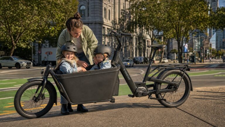 Mother helping two helmeted children seated in a black Trek electric cargo bike on a sunny urban bike lane